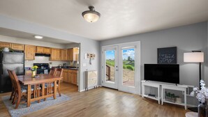 Una cocina moderna con armarios de madera, un comedor con mesa y sillas de madera, un televisor sobre un mueble bajo y vistas a través de puertas de vidrio.