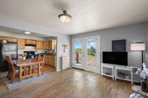 Eine moderne Küche mit Holzausstattung, eine Essecke mit Holztisch und -stühlen, ein Fernseher auf einem Schrank und Blick durch Glastüren.