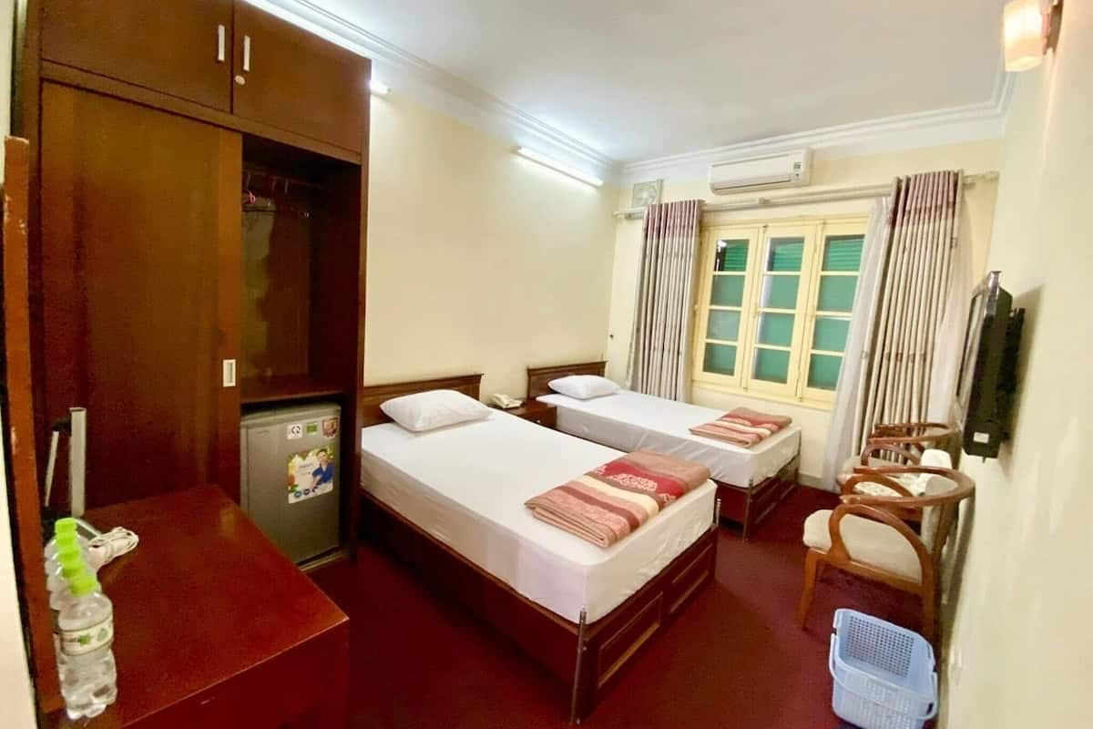 Quarto de hotel com duas camas, uma mesa, frigorífico, televisão e janela com cortinas.