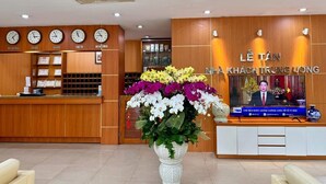 A reception area with a wooden reception desk, a large floral arrangement, clocks on the wall, and a television screen displaying a news broadcast. - Nha Khach TW (Hanoi)