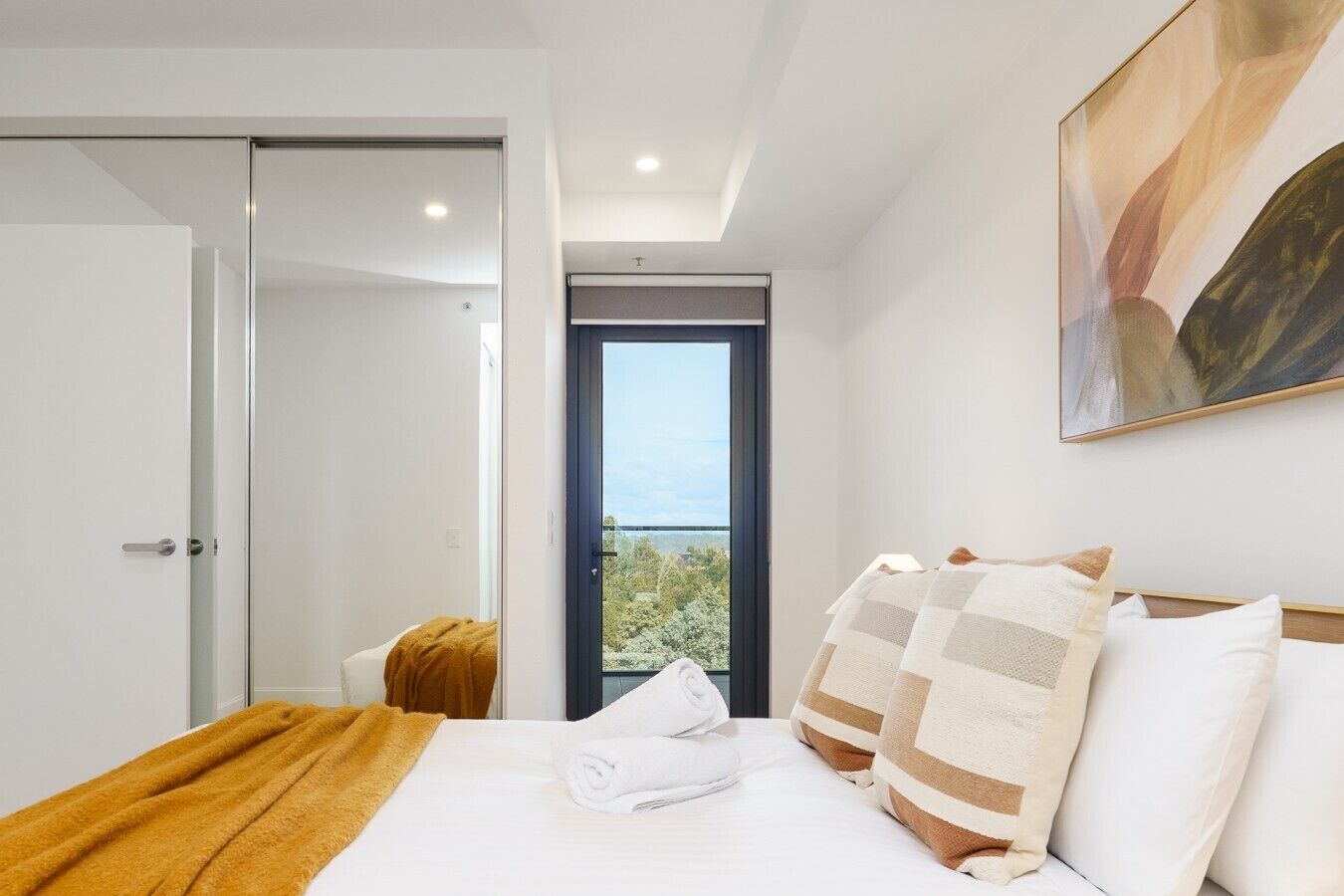 A hotel room with a bed, a window with a view, a painting on the wall, and a door leading to another room.