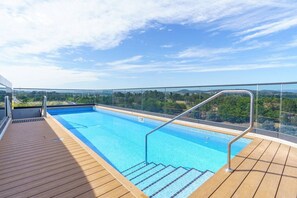 Ein Schwimmbecken mit Holzterrasse und Glasgeländer, umgeben von einer reizvollen Aussicht.