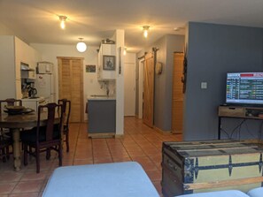 A room with a bed, a wooden chest, a dining area with chairs, a kitchen, and a television displaying a menu.