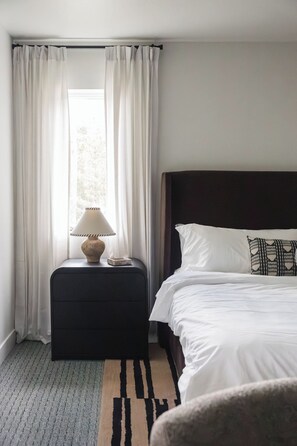 A hotel room with a bed, a bedside table with a lamp, a rug, and sheer curtains.
