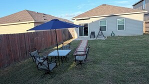 Ein Garten mit einer Terrasse, einem Tisch und Stühlen, einem Holzzaun und einem Haus mit Garage.
