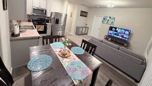 Eine moderne Küche mit Edelstahlgeräten, ein Essbereich mit einem Holztisch und blauen Tischsets sowie ein Wohnbereich mit einem grauen Sofa und einem Flachbildfernseher.