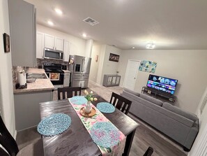Una cocina moderna con electrodomésticos de acero inoxidable, un comedor con mesa de madera y individuales azules, y una sala de estar con sofá gris y televisor de pantalla plana.