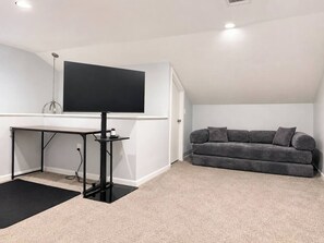 Ein Zimmer mit Schreibtisch, schwarzem Stuhl, grauem Sofa und einem großen, schwarzen Flachbildfernseher an der Wand.