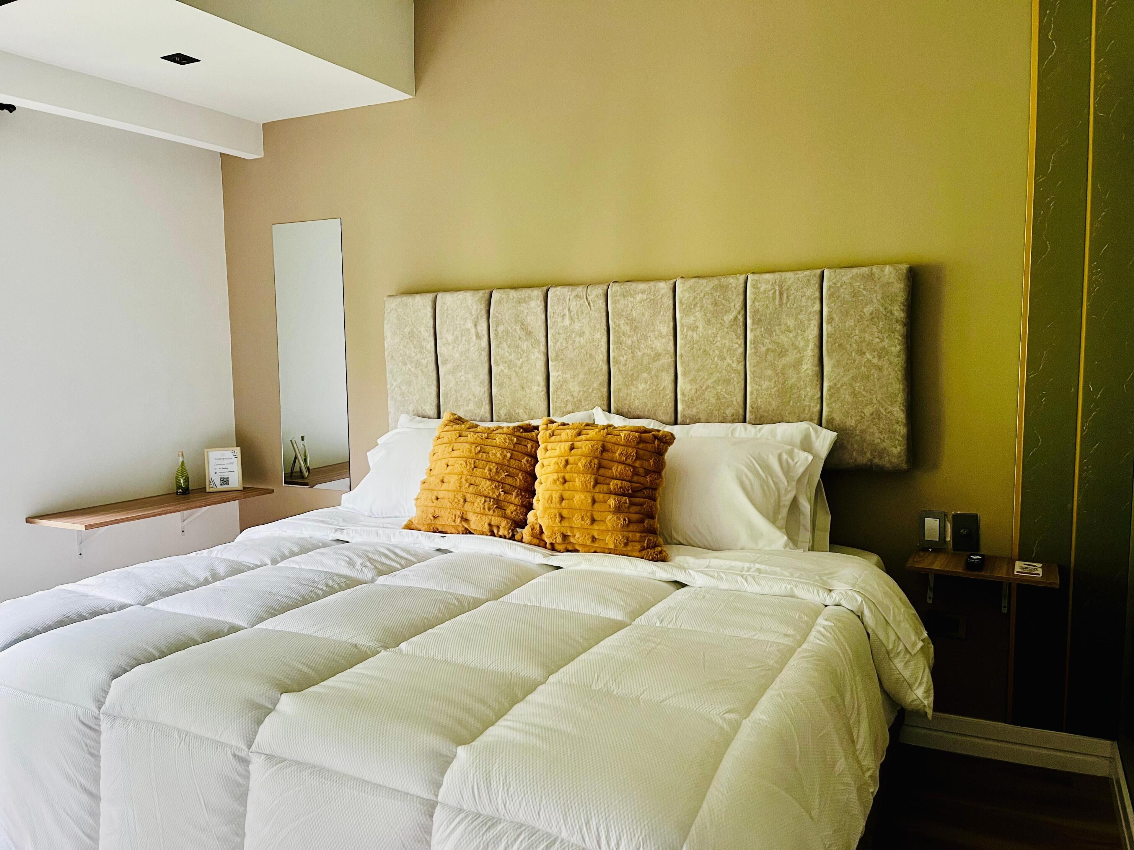 Una habitación de hotel con cama, cabecera, mesita de noche y espejo.