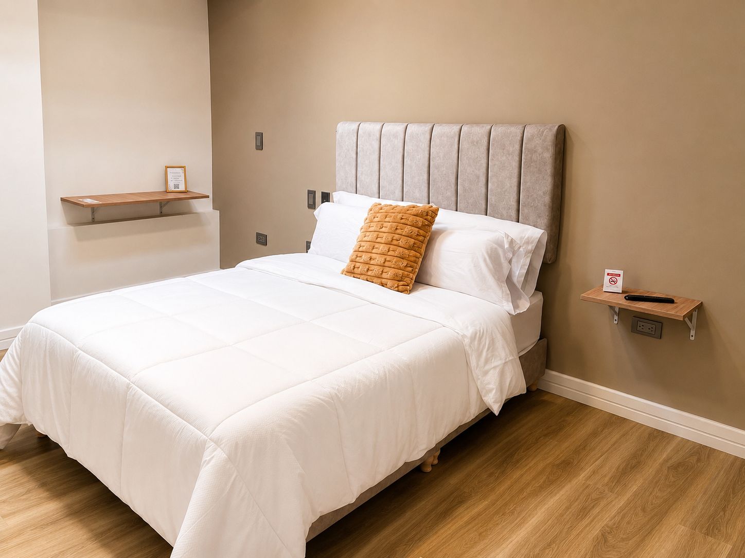 Una cama bien hecha con sábanas blancas y una única almohada decorativa, un pequeño estante con un cartel y un enchufe en la pared.