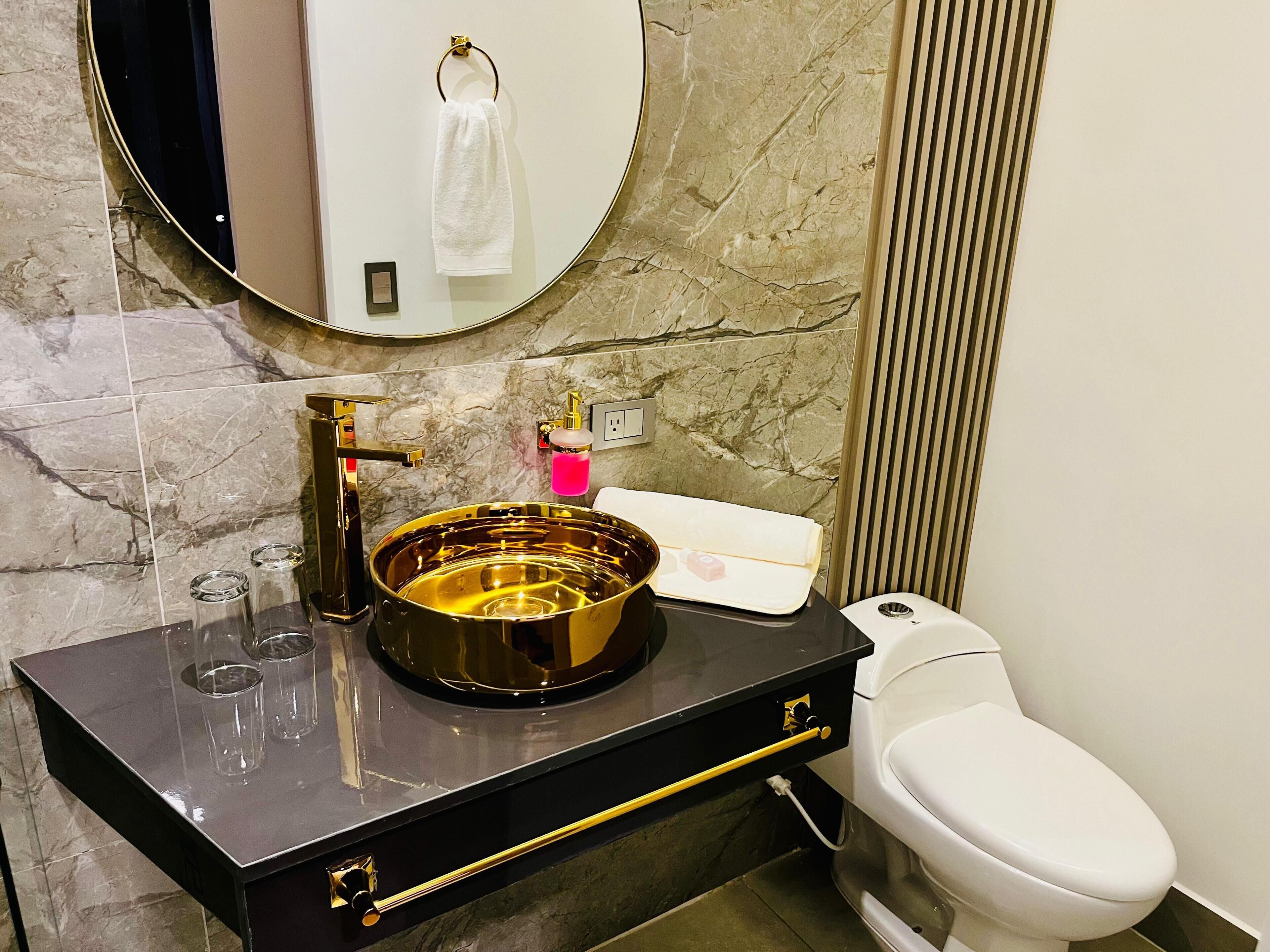 A bathroom with a black vanity, a gold sink, a white toilet, and a marble backsplash.