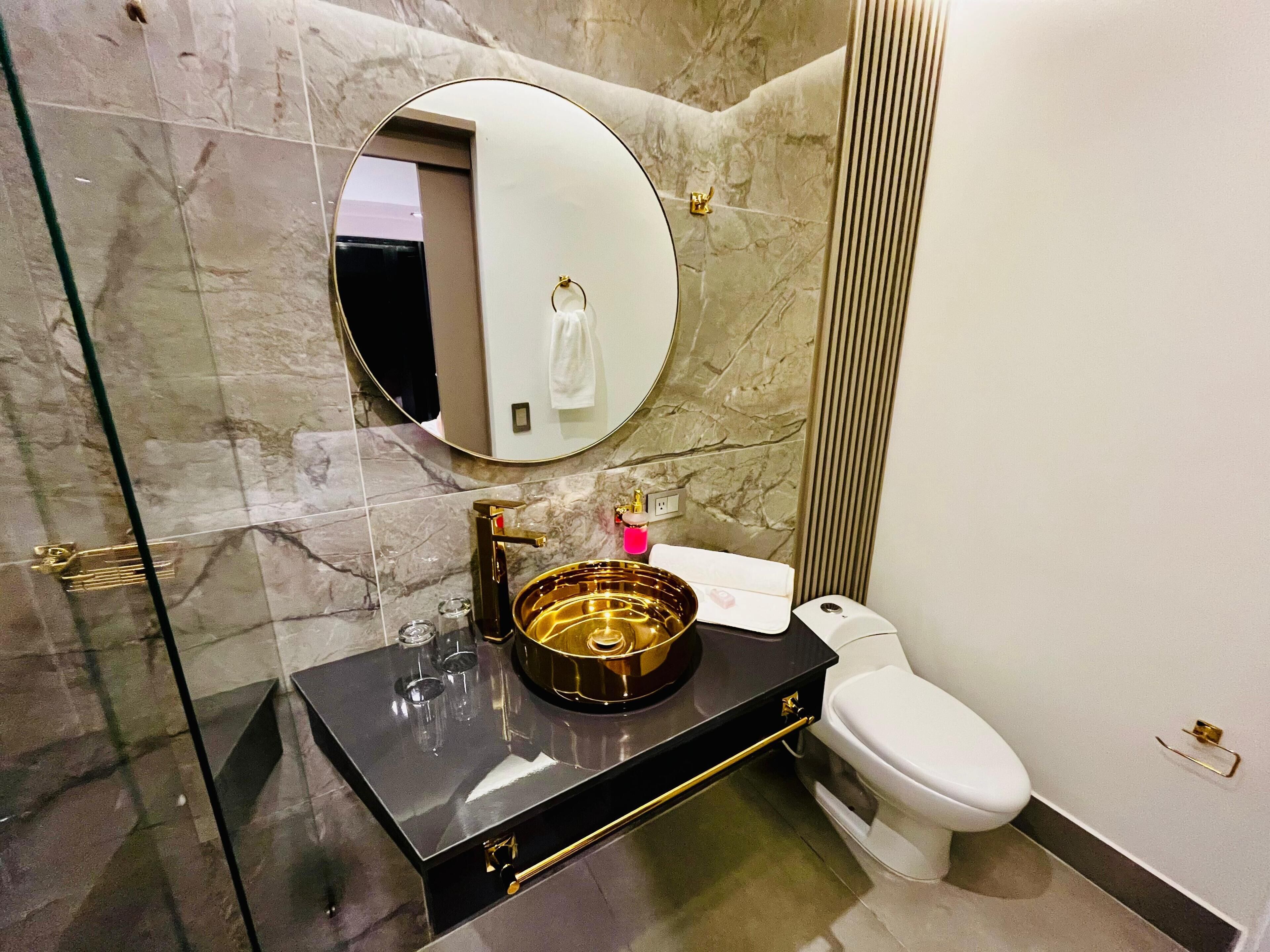A bathroom with a marble wall, a round mirror, a gold sink, and a white toilet.