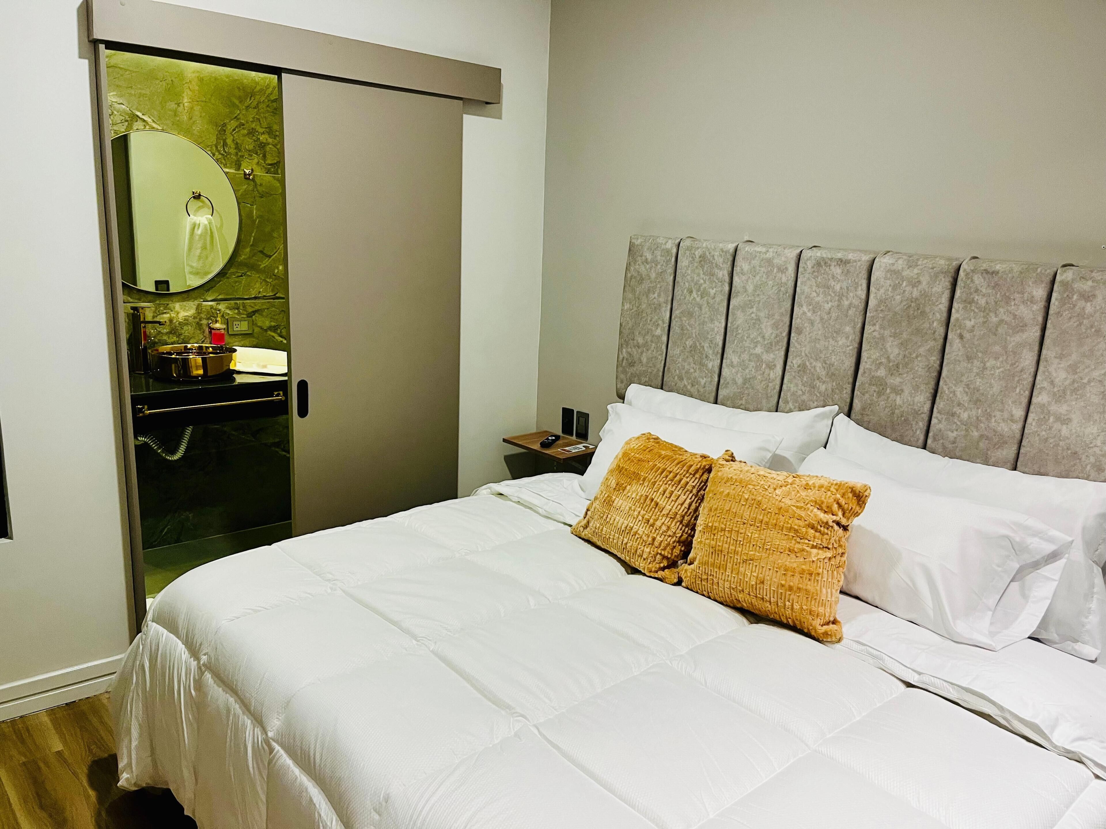 A hotel room with a bed, a nightstand, a mirror, and a bathroom with a sink and toiletries.