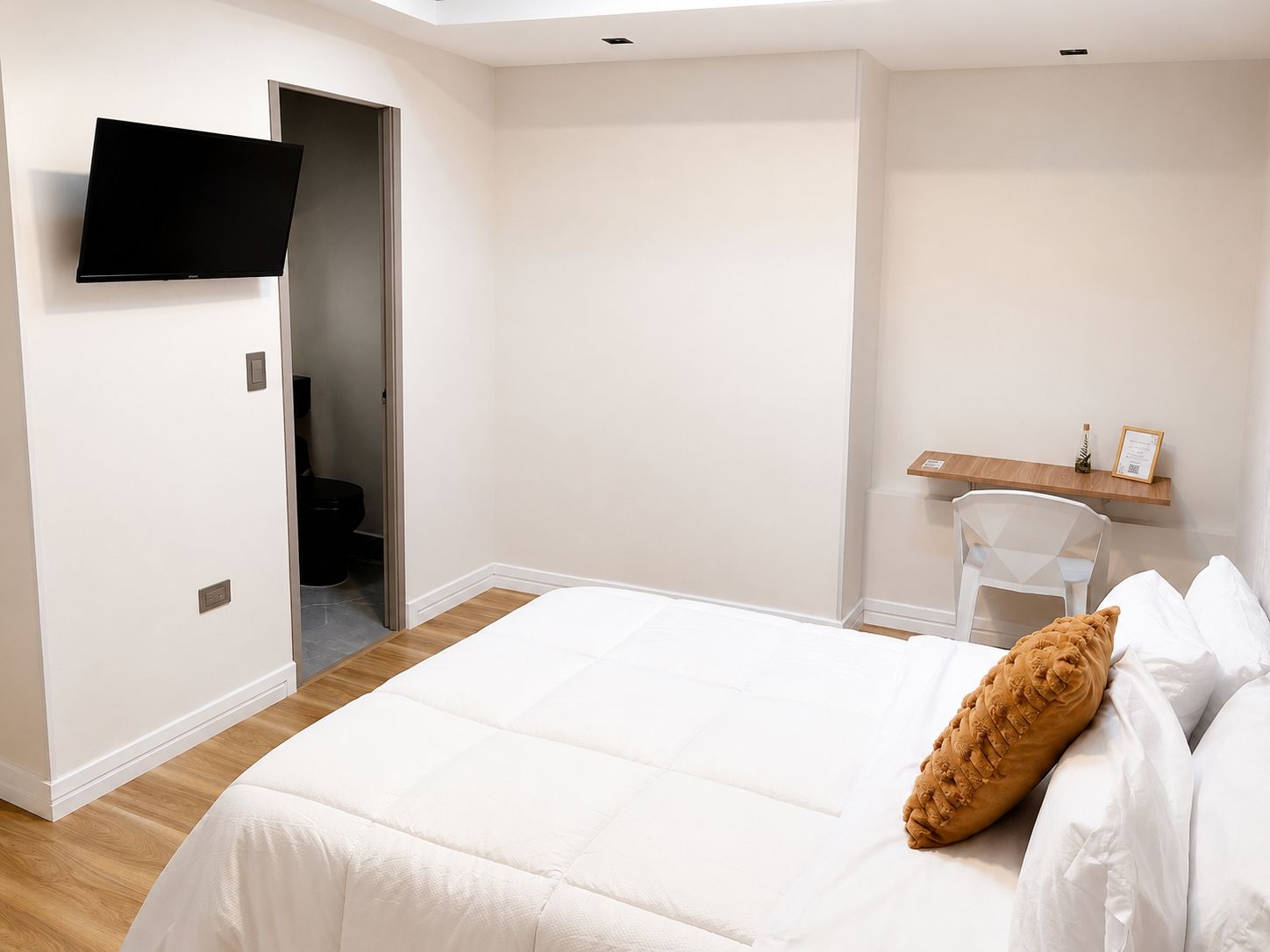 Un dormitorio con cama, televisor montado en la pared, una silla de madera y un pequeño estante con objetos.