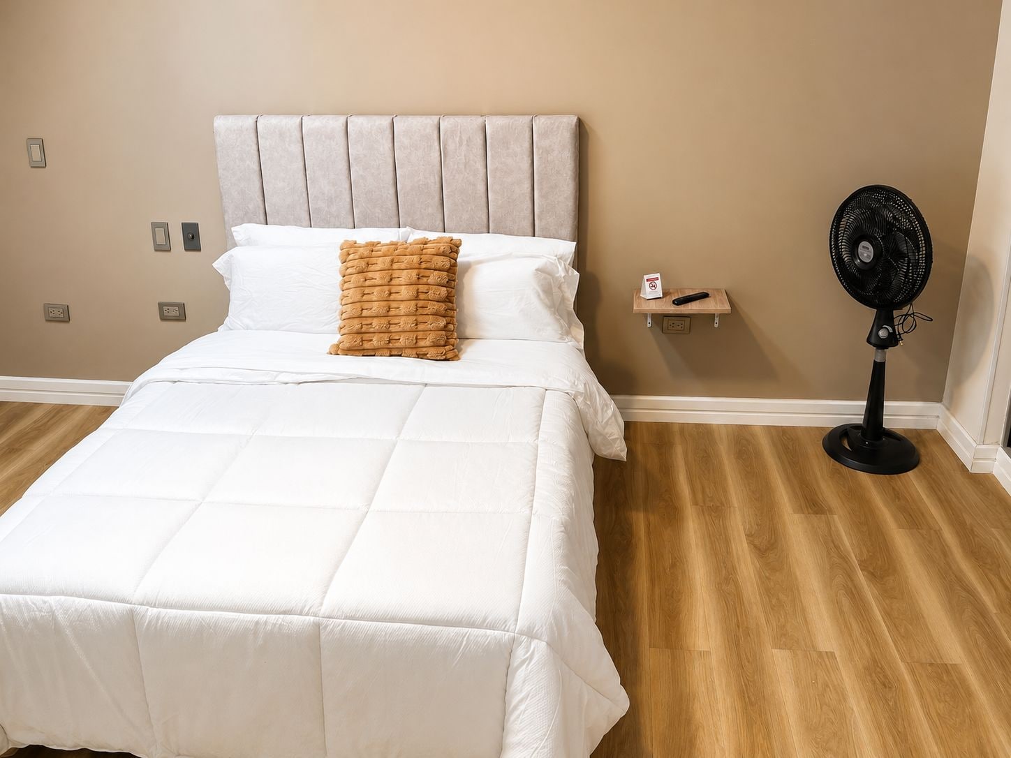 Un lit bien fait, avec une couette blanche et un oreiller marron à texture intéressante, une table de chevet avec une télécommande et un ventilateur sur pied.