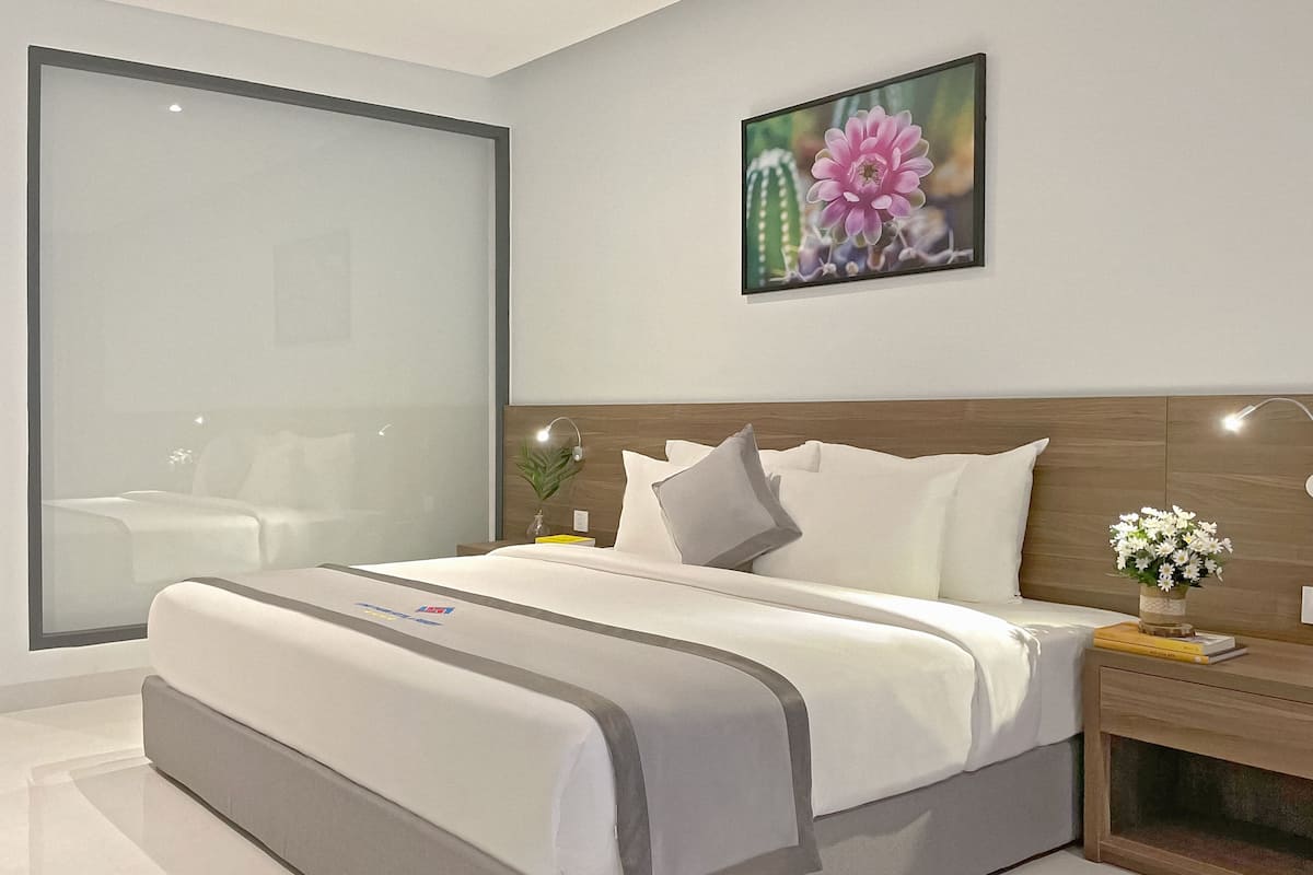 A modern hotel room with a large bed, bedside tables, a nightstand, a wall-mounted lamp, and a large window.