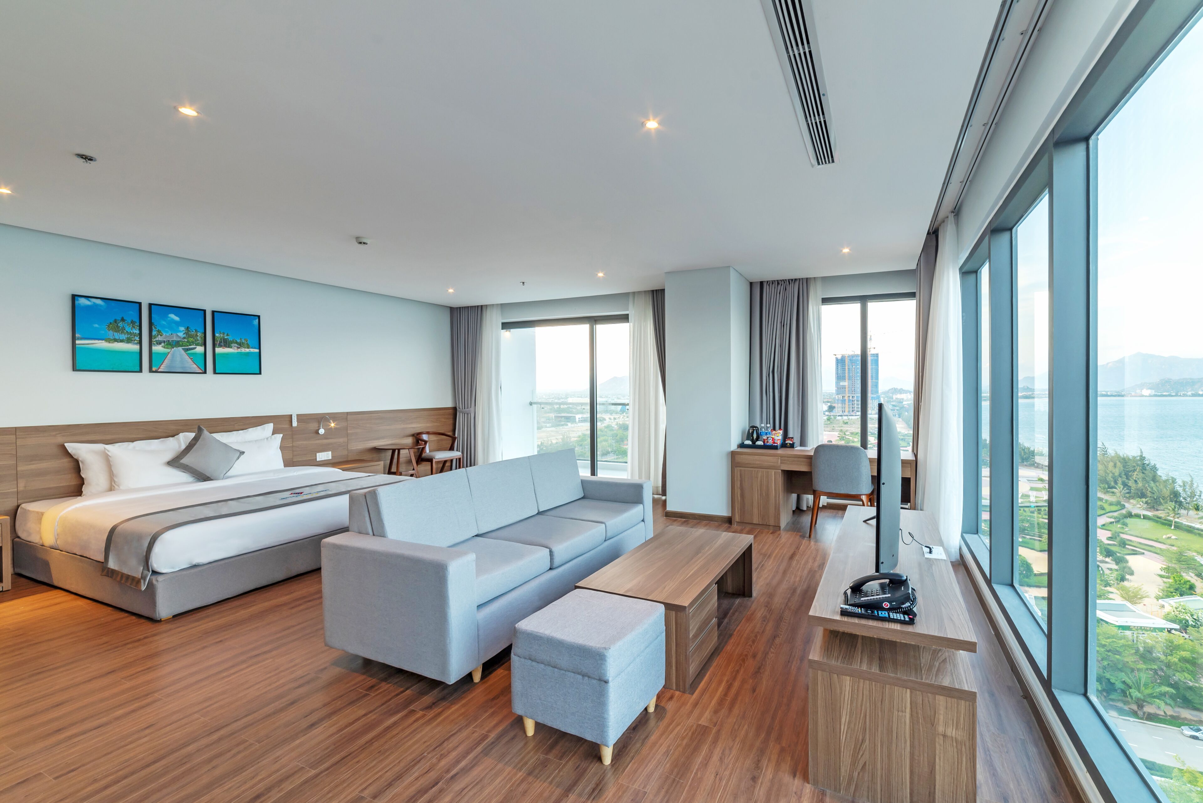 A modern hotel room with a bed, sofa, desk, and large windows offering a city view.