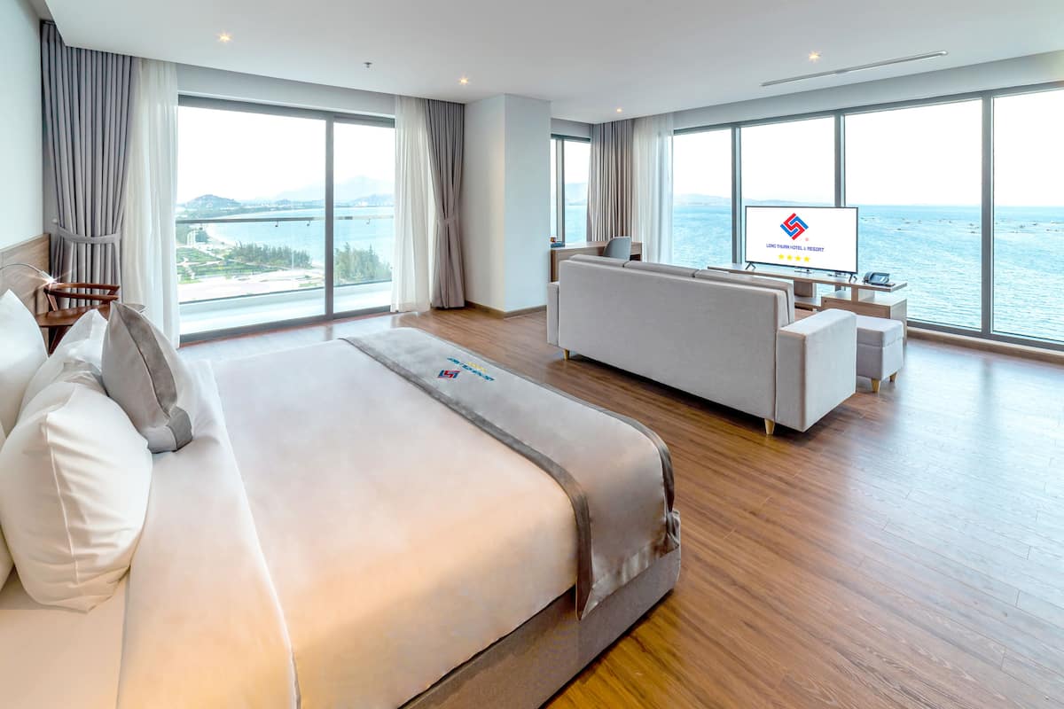 A modern hotel room with a large bed, a sofa, a TV, and a view of the sea.