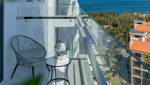 A modern balcony with a glass railing, a wicker chair, a small table, and a view of the ocean and surrounding buildings. - Long Thuan Hotel & Resort (Dong Hai)
