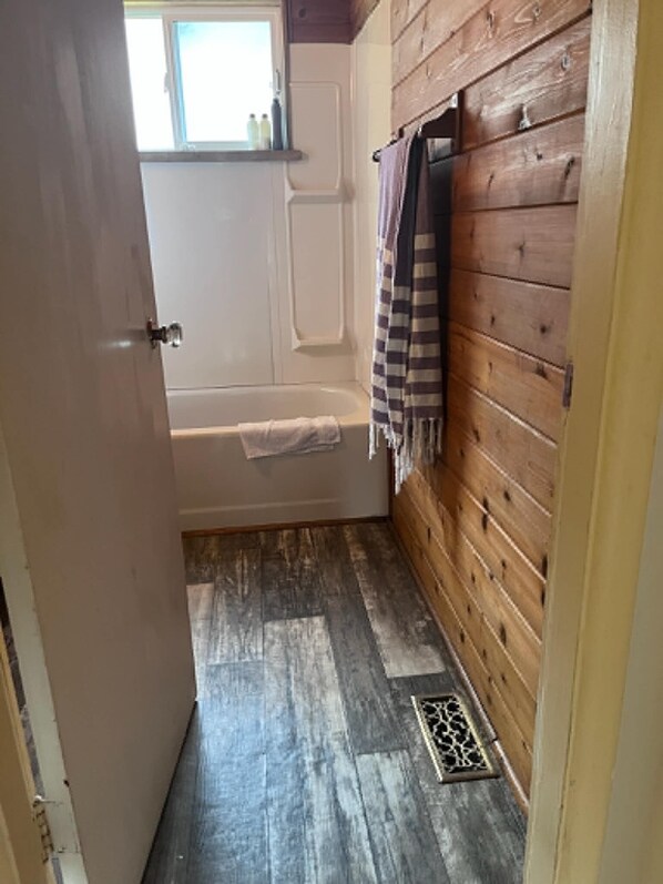 A narrow bathroom with a wooden wall, a bathtub, and a window.