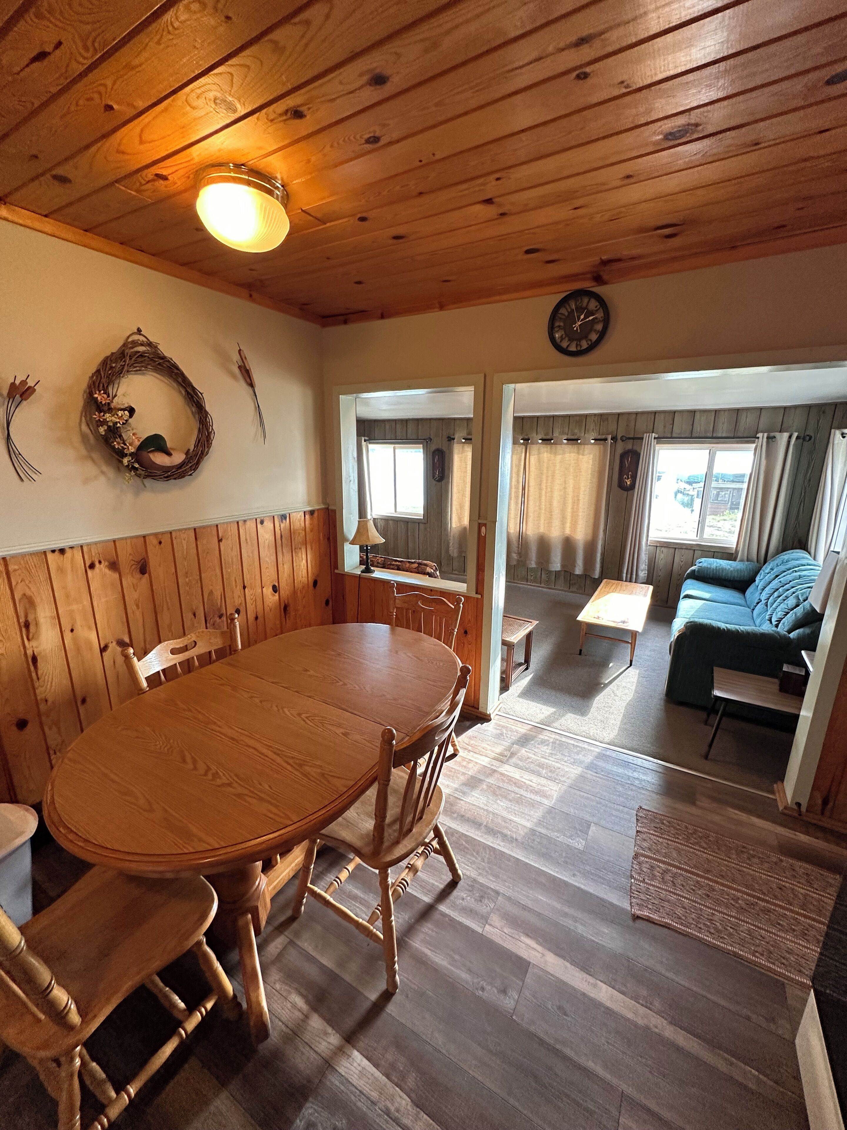 A wooden dining area with a table and chairs, a clock on the wall, and a view into the living room through a doorway.