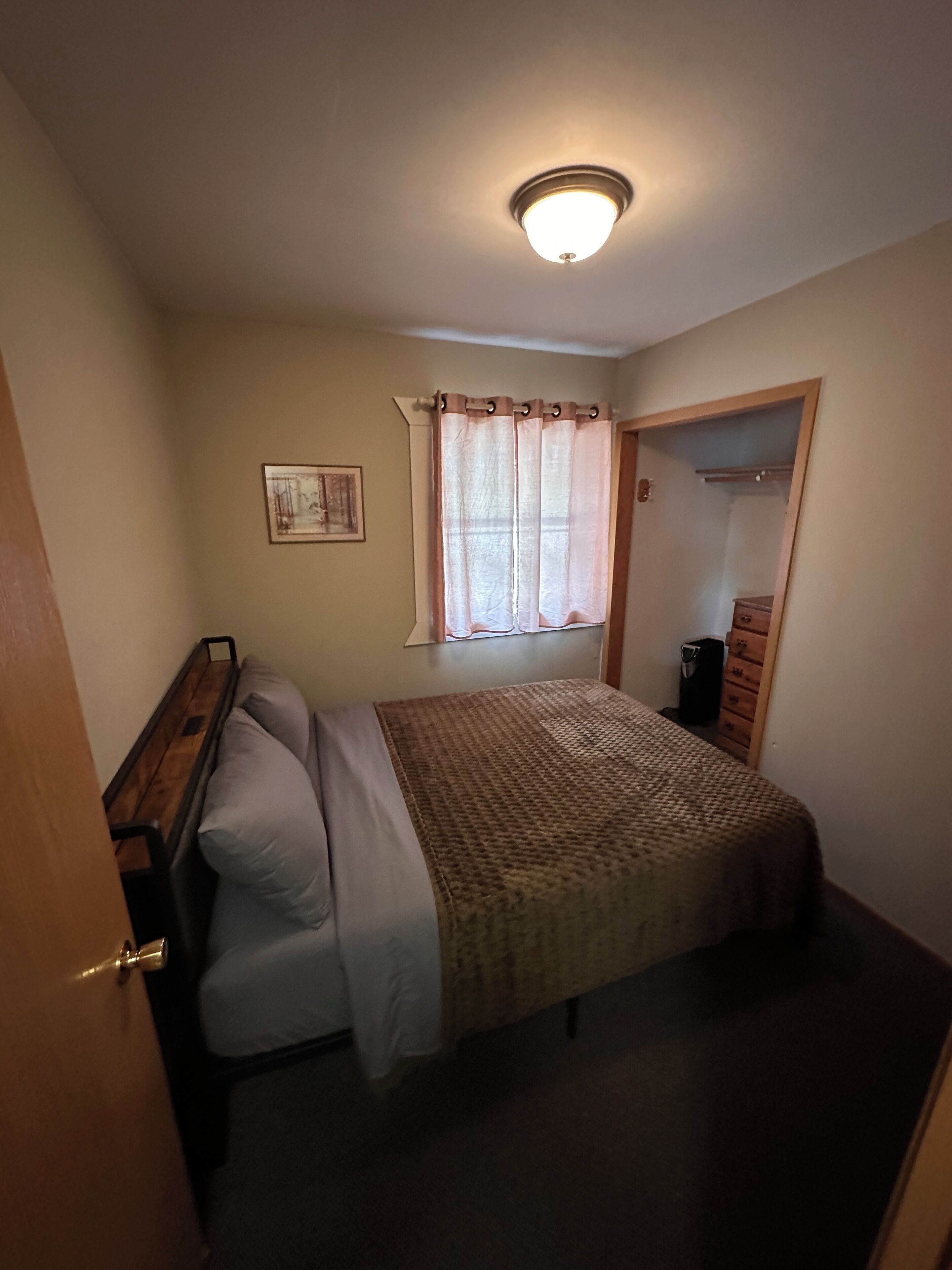 A bedroom with a bed, a window with curtains, a door, and a ceiling light.