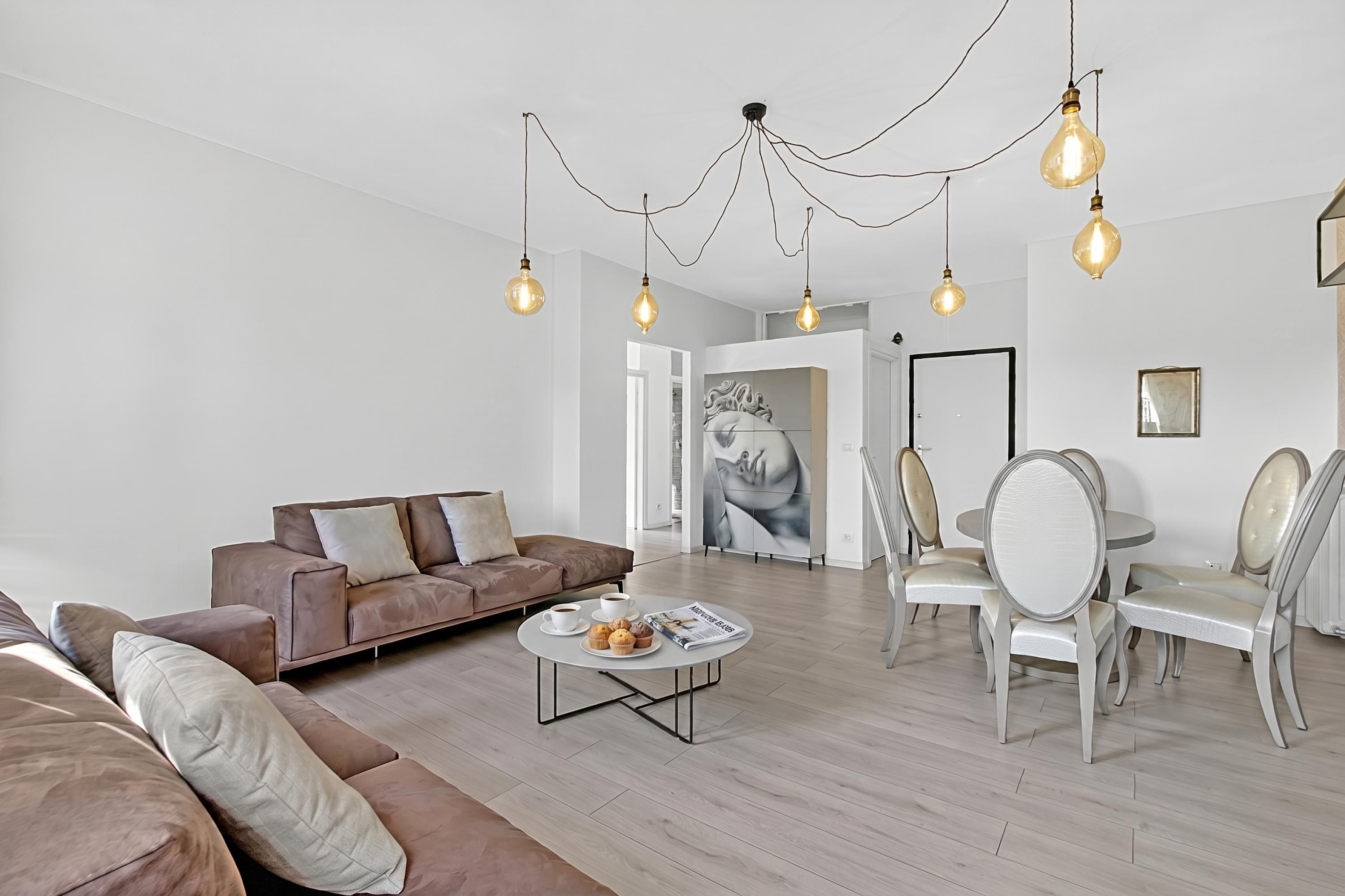 Un salon moderne avec un canapé marron, une table basse ronde sur laquelle sont disposées des pâtisseries, et un coin repas avec des chaises blanches.