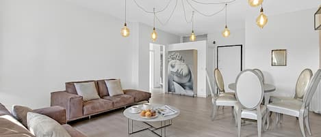 Un salon moderne avec un canapé marron, une table basse ronde sur laquelle sont disposées des pâtisseries, et un coin repas avec des chaises blanches.