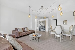 A modern living room with a brown sofa, a round coffee table with pastries, and a dining area with white chairs. - Bono Deluxe: 3BR Home in Desenzano (Desenzano del Garda)