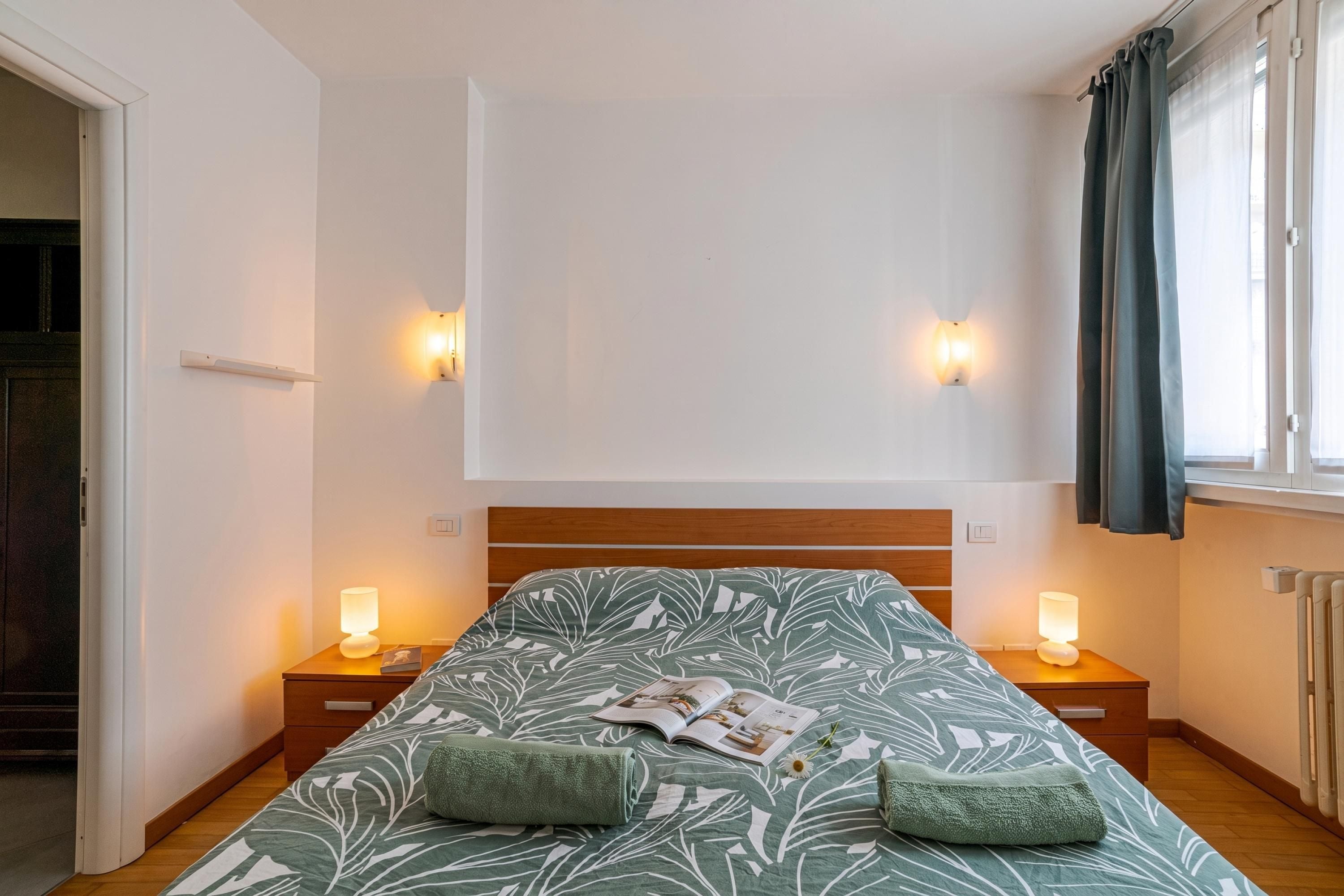A bedroom with a bed, two bedside tables with lamps, a window with curtains, and a wall-mounted light fixture.