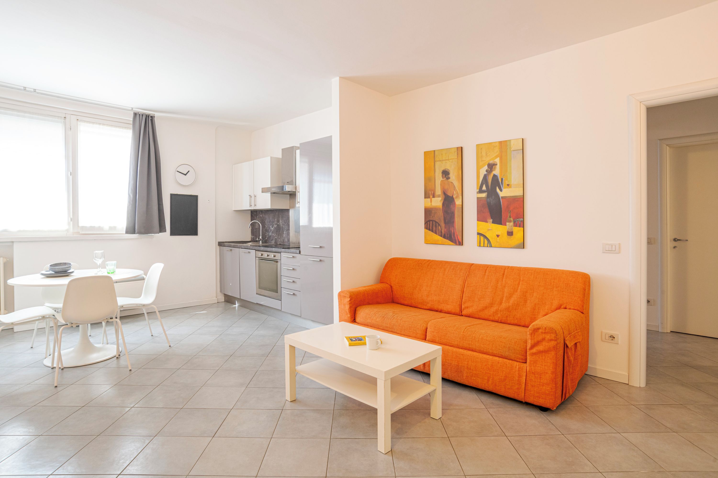 Una sala moderna con una mesa de centro blanca, un sofá naranja, un comedor con sillas blancas y una cocina con gabinetes blancos.