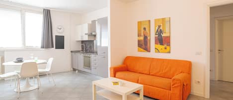 Una sala moderna con una mesa de centro blanca, un sofá naranja, un comedor con sillas blancas y una cocina con gabinetes blancos.