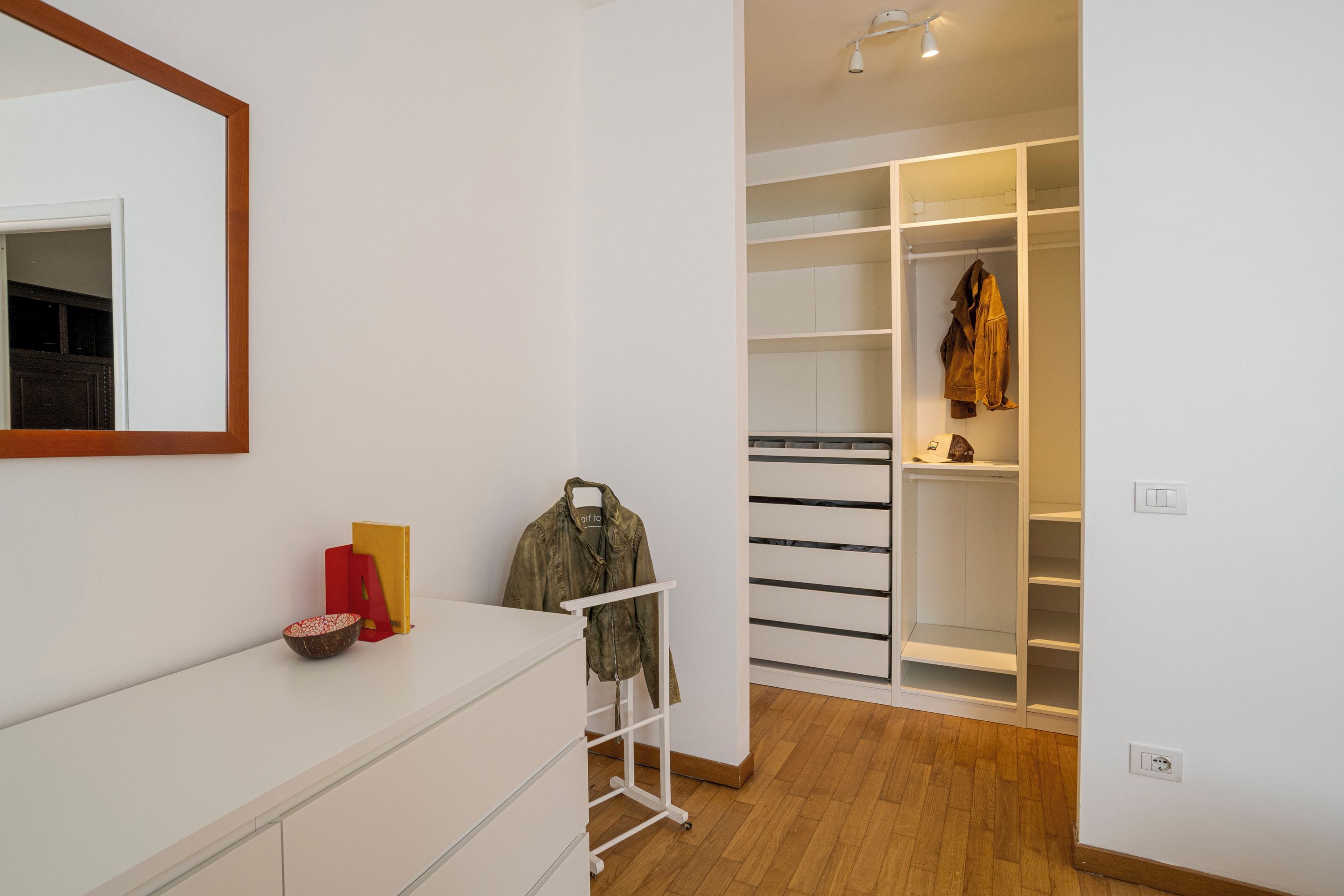 A modern room with a white dresser, a mirror, a coat rack, and a walk-in closet with shelves.