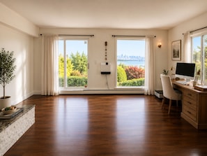 Ein geräumiges Zimmer mit Holzboden, einem Schreibtisch mit Computer, einem Stuhl und Blick auf eine Stadt durch große Fenster.