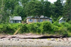 A coastal house with a wooden deck, surrounded by dense greenery and a rocky shoreline.