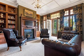 A room with a fireplace, leather armchairs, a sofa, a bookshelf, and a large window with curtains. - Earnscleugh Castle (Alexandra)