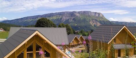 A row of wooden cabins with sloped roofs, set against a backdrop of mountains and a clear sky.