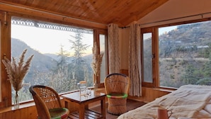 Una baita in legno con un tavolo, due sedie in vimini, un letto e ampie finestre che offrono vista sulle montagne.