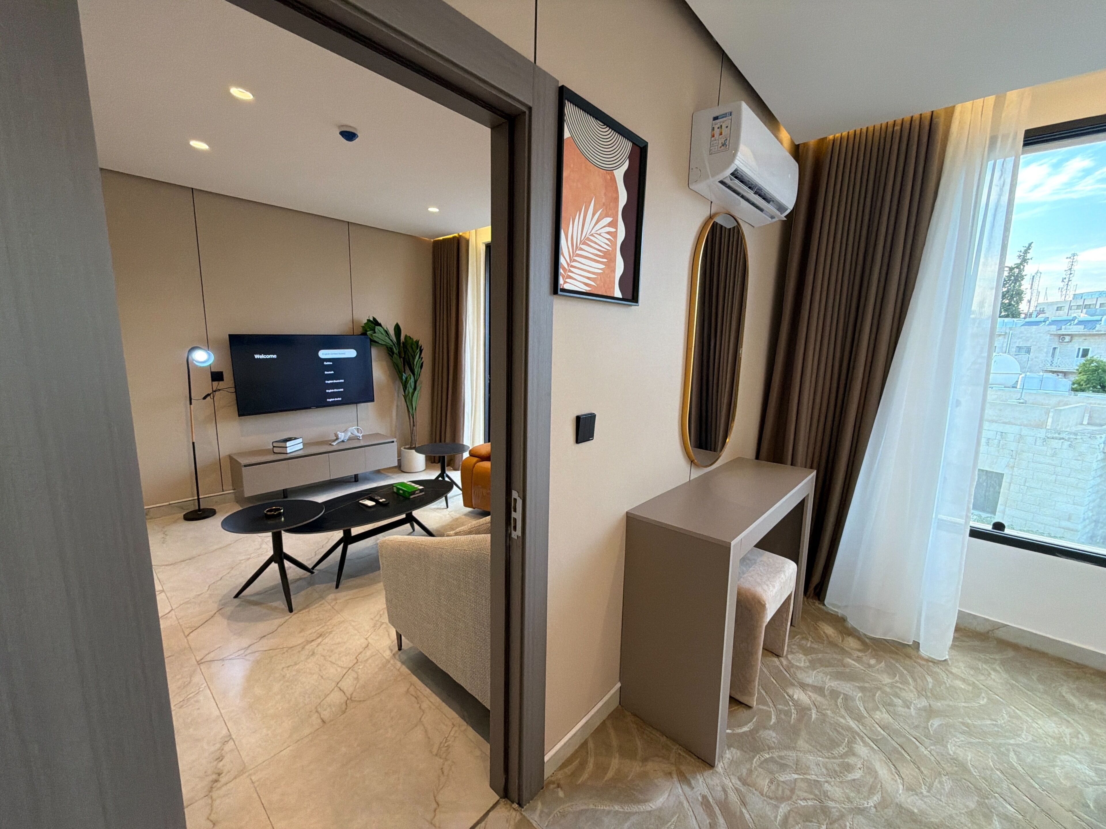 A modern hotel room with a flat-screen TV, a desk, a chair, a small table, and a potted plant.