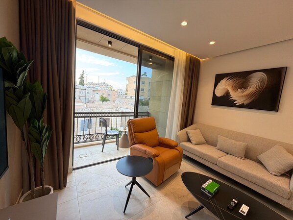 Ein modernes Wohnzimmer mit einer Couch, einem Sessel und einem kleinen Tisch, einem großen Fenster mit Blick auf Gebäude und einem Gemälde an der Wand.