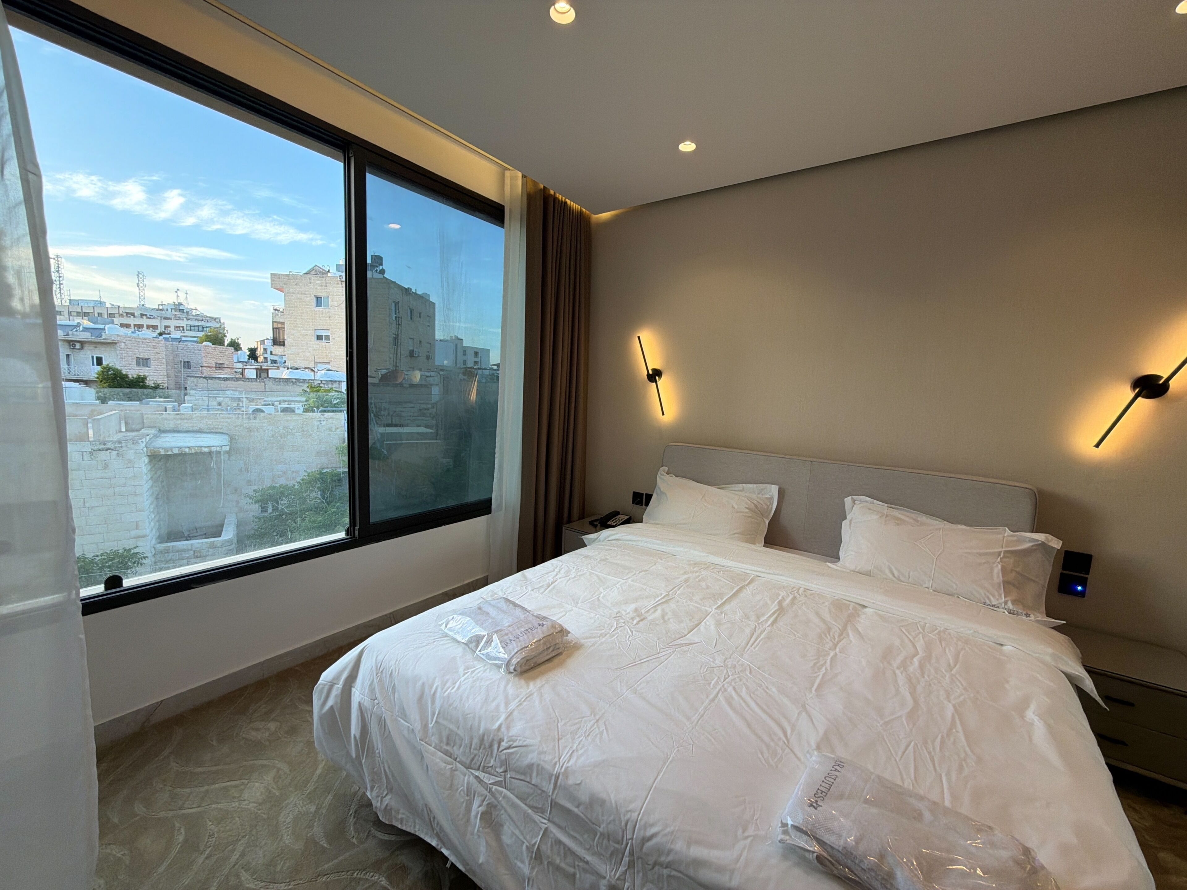 A hotel room with a large window, a bed with white bedding, a nightstand, and a wall-mounted light fixture.