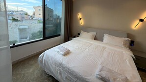 A hotel room with a large window, a bed with white bedding, a nightstand, and a wall-mounted light fixture. - 412 Tara-jre Jabal Al Lweibdeh With Stunning View (Amman)