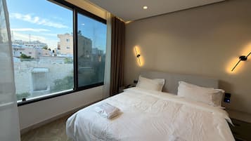 A hotel room with a large window, a bed with white bedding, a nightstand, and a wall-mounted light fixture.