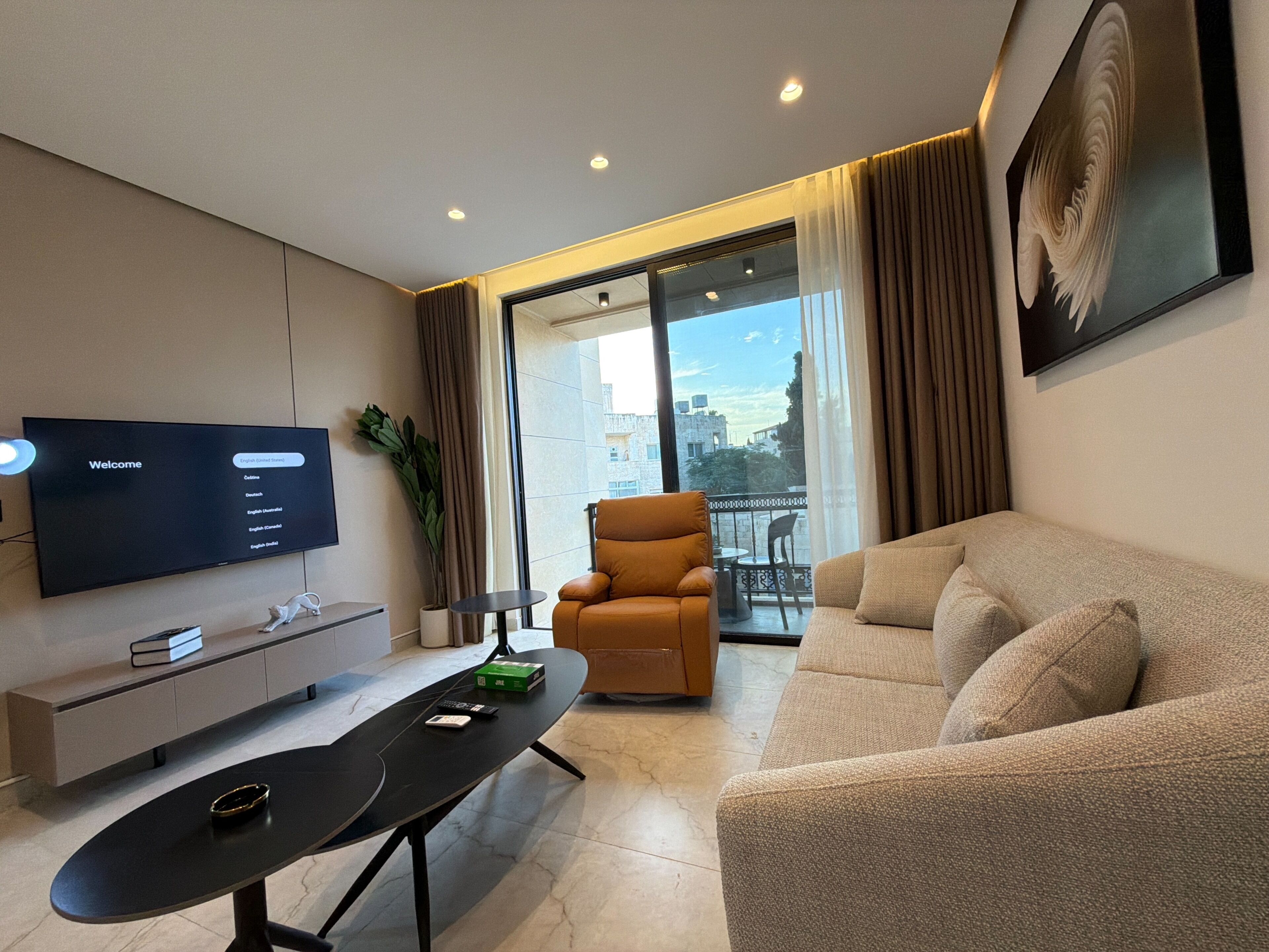 A modern living room with a flat-screen TV, a sofa, a chair, a small table, and a balcony with a view.