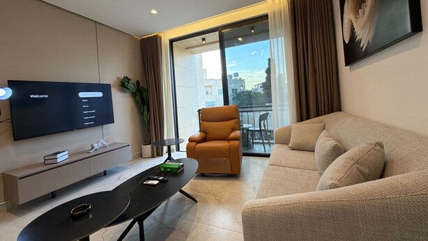 A modern living room with a flat-screen TV, a sofa, a chair, a small table, and a balcony with a view. - 412 Tara-jre Jabal Al Lweibdeh With Stunning View (Amman)