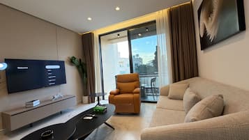 A modern living room with a flat-screen TV, a sofa, a chair, a small table, and a balcony with a view.