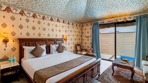 A hotel room with a large bed, a wooden headboard, a nightstand with a lamp, a small table with a vase, and a view of the outside through a sliding door. - Bijainagar Fort (Bijaynagar)