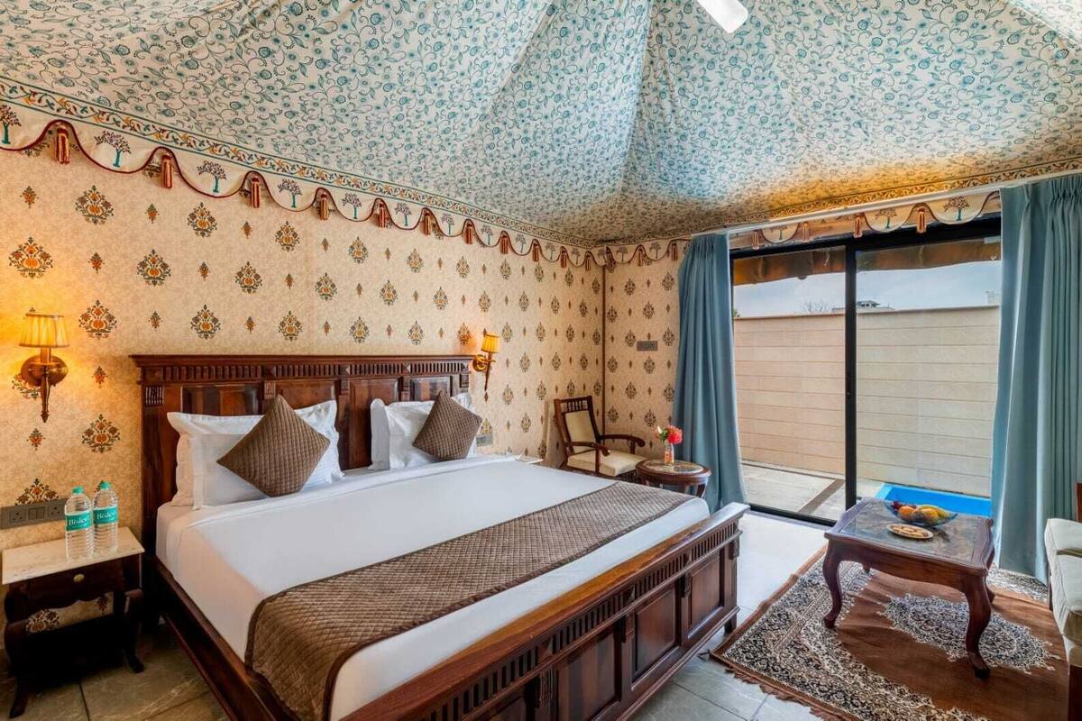 Habitación de hotel con una cama grande, cabecera de madera, mesita de noche con lámpara, pequeña mesa con un florero y vistas al exterior a través de una puerta corrediza.