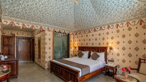 A hotel room with a large bed, a desk, a chair, a small table with fruit, and patterned wallpaper. - Bijainagar Fort (Bijaynagar)