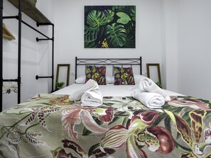 Ein ordentlich bezogenes Bett mit floraler Bettwäsche, Wandkunst im tropischen Stil und einem schwarzen Metall-Kopfteil.